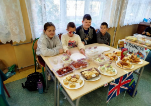 Grupa uczniów klasy 5 prowadzi stoisko kulinarne poświęcone Wielkiej Brytanii. Na stole znajdują się brytyjskie przysmaki, w tym kanapki, ciasta i inne przekąski, które są udekorowane małymi flagami Union Jack. Stół jest również ozdobiony dużą flagą Wielkiej Brytanii.