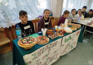 Klasa 6 prowadzi stoisko kulinarne poświęcone Włochom. Na stole znajdują się pizze, tiramisu, spaghetti, koreczki oliwkowe i pieczywo. Przed stołem wiszą plakaty z napisami ‘Zapraszamy u nas smaczniej’ i ‘Włochy’, ozdobione włoską flagą i wieżą w Pizie.