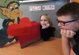 czerwona skrzynka pocztowa z serduszkiem, której przyglądają się Majka i Filip- zaangażowani w przygotowanie szkolnych walentynek przedstawiciele SU