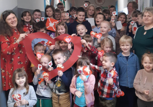 zdjęcie roześmianych wszystkich dzieci z serduszkowymi lizakami od SU.Nad nimi widać czerwone, uśmiechnięte serce, które trzyma Filip, w pierwszym szeregu buzie przedszkolaków w wyciętym konturze serca, które trzyma p. Kierownik.