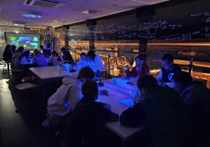 Grupa uczniów siedzi przy stolikach w laboratorium, przy których świecą lampy UV.