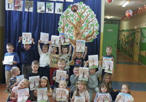 Zdjęcie grupowe dzieci z grupy Zuchy na tle dekoracji Dzień Drzewa, dzieci prezentują swoje prace - drzewo malowane kredkami.