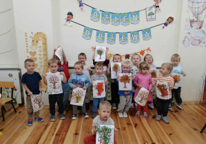 Zdjęcie grupowe w sali dzieci z grupy Kamyczki, przedszkolaki trzymają w dłoniach drzewa malowane farbami plakatowymi.
