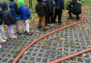 Przedszkolaki ustawione w rzędzie oraz strażacy przygotowujący stanowisko do obsługi węża gaśniczego.