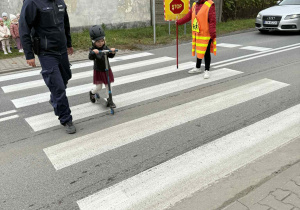 Dziewczynka z grupy Słoneczka przeprowadza hulajnogę pod nadzorem policjanta.