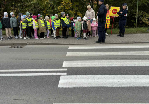 Grupa dzieci stoi przy przejściu dla pieszych i słucha policjantów.