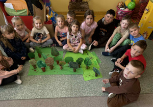 Dziewczynki siedzą na podłodze a przed nimi stoją drzewa z recyklingu.