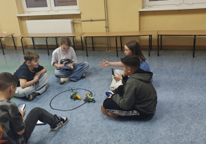 Uczniowie projektują i prezentują swoje sumoboty. Siedzą w kręgu na korytarzu szkolnym.