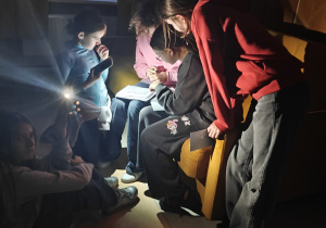 Uczennice czytają legendę o zniszczeniu Biblioteki Aleksandryjskiej przy włączonej latarce w telefonie. Znajdują się na korytarzu szkolnym.