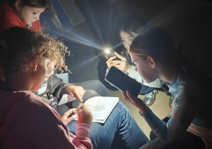 Uczennice pracują nad odczytaniem hasła-polecenia według podanego kodu. Jest ciemno. Mają włączoną latarkę w telefonie.