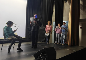 Uczniowie grają role Hadesa i Zeusa w scence na podstawie mitu ,,Demeter i Kora” na scenie sali widowiskowej GOK. Z boku sceny stoją pozostali bohaterowie występujący w scence.