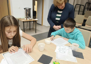 Uczniowie rozwiązują krzyżówki i wykreślanki. Obok uczniów stoi pani polonistka.