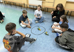 Uczniowie prezentują wyniki swojej pracy robotycznej przy pani bibliotekarce. Wszyscy znajdują się na korytarzu szkolnym.