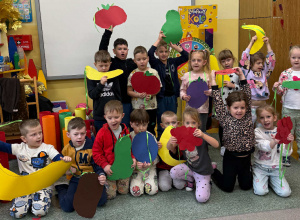 Dzieci z grupy Żabki pozują do grupowego zdjęcia z dużymi owocami i warzywami z papieru.