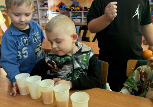 Dzieci porównują kto ma najwięcej soku pomarańczowego w kubeczku a kto najmniej.