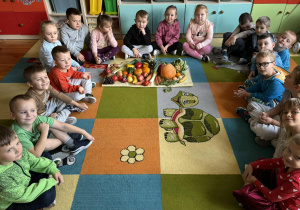 Dzieci w półkolu z grupy Promyczki prezentują przyniesione owoce i warzywa.