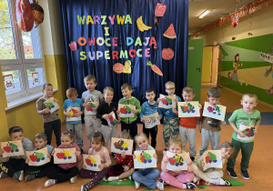 Przedszkolaki z grupy Promyczki prezentują swoją prace techniczną na tle dekoracji z okazji Dnia Owoców i Warzyw.
