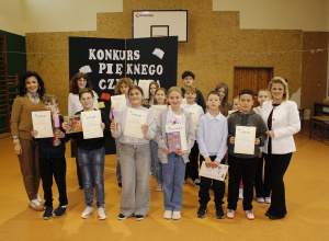 Pamiątkowe zdjęcie Pani Dyrektor, nauczycieli i uczniów na zakończenie Konkursu. Uczniowie trzymają nagrody i dyplomy. Z tyłu tablica dekoracyjna przygotowana na ,,Konkurs Pięknego Czytania”