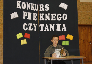 Uczeń klasy VI czyta fragment powieści pt. ,,Opowieści z Narnii. Lew, czarownica i stara szafa”. Siedzi przy stoliku. Za nim znajduje się tablica dekoracyjna z hasłem ,,Konkurs Pięknego Czytania”.