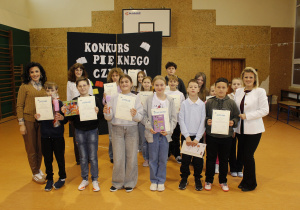 Pamiątkowe zdjęcie Pani Dyrektor, nauczycieli i uczniów na zakończenie Konkursu. Uczniowie trzymają nagrody i dyplomy. Z tyłu tablica dekoracyjna przygotowana na ,,Konkurs Pięknego Czytania”.