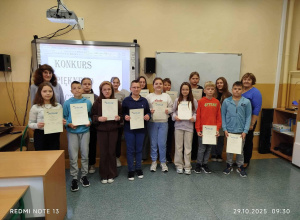Uczniowie stoją w klasie przed tablicami. W rękach trzymają dyplomy. Razem z uczniami stoi Pani bibliotekarka i Pani polonistka.