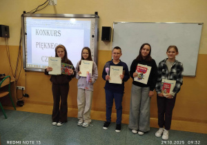 Uczniowie nagrodzeni w ,,Konkursie Pięknego Czytania” stoją w klasie i trzymają nagrody i dyplomy. Za nimi na jednej z tablic znajduje się hasło ,,Konkurs Pięknego Czytania”.