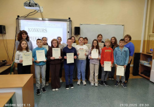 Uczniowie stoją w klasie przed tablicami. W rękach trzymają dyplomy. Razem z uczniami stoi Pani bibliotekarka i Pani polonistka.