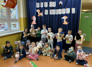 Zdjęcie grupowe dzieci z grupy Promyczki w Dniu Kudelka, przedszkolaki trzymają w dłoniach swoje maskotki bądź figurki piesków oraz prace techniczną wykonaną techniką origami.