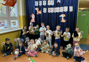 Zdjęcie grupowe dzieci z grupy Promyczki w Dniu Kudelka, przedszkolaki trzymają w dłoniach swoje maskotki bądź figurki piesków oraz prace techniczną wykonaną techniką origami.