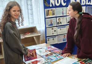 Uczennice klasy siódmej oglądają książki w bibliotece szkolnej w Rzeczycy w czasie Kiermaszu Książki. Za uczennicami znajduje się wystawka książek ,,Dlaczego ludzie sławni czytają więcej?”