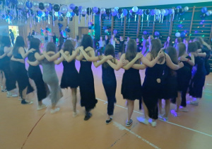 Uczennice tańczą w kole na sali trzymając się za szyje.