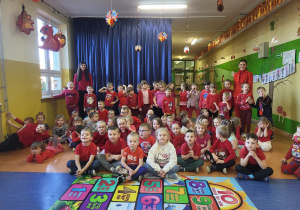 Przedszkolaki wraz z psychologiem i Panią Dyrektor ubrani są na czerwono część grupy siedzi na podłodze część stoi na korytarzu w przedszkolu.