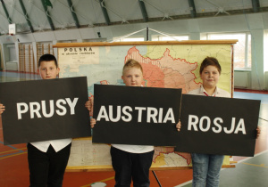 Trzech uczniów na tle mapy Polski w okresie rozbiorów, trzymających tabliczki z nazwami zaborców.