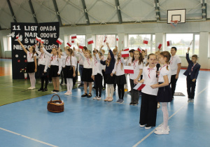 Dwie uczennice śpiewające utwór patriotyczny, pozostali uczniowie trzymają w górze chorągiewki.