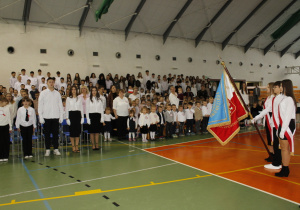 Uczniowie śpiewający hymn państwowy, po prawej stronie poczet sztandarowy.