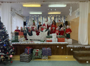 Grupa Żabki z Mikołajem, Panią Dyrektor Agatą Ewą Ostalską , Panią Zastępcą Wójta Gminy Rzeczyca Iwoną Łuszcz- Krawczyk i Panią Kierownik Bernadetą Grot, na tle dekoracji zimowej z saniami.