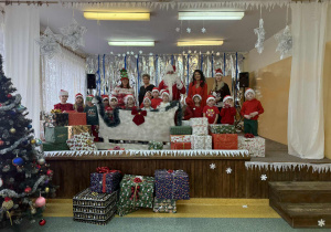 Grupa Żabki z Mikołajem, Panią Dyrektor Agatą Ewą Ostalską , Panią Zastępcą Wójta Gminy Rzeczyca Iwoną Łuszcz- Krawczyk i Panią Kierownik Bernadetą Grot, na tle dekoracji zimowej z saniami.