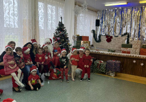 Grupa Słoneczka z Mikołajem, Panią Dyrektor Agata Ewą Ostalską, Panią Zastępcą Wójta Gminy Rzeczyca Iwona Łuszcz- Krawczyk i Panią Kierownik Bernadetą Grot, na tle dekoracji zimowej z saniami.