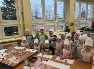 Dzieci z grupy Żabki w białych strojach kucharskich na tle okien sali przedszkolnej.