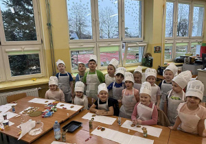 Dzieci z grupy Żabki w białych strojach kucharskich na tle okien sali przedszkolnej.