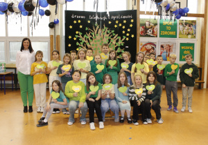 Grupowe zdjęcie uczniów klasy 4 na tle drzewa tolerancji i życzliwości. Uczniowie trzymają w rękach serduszka wykonane z papieru.