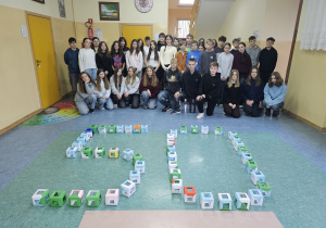 Uczniowie klas siódmych na szkolnym korytarzu. Przed uczniami ułożona jest liczba 50 z pięćdziesięciu sześcianów modułowych.