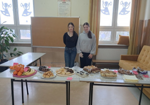 Uczennice klasy VIII b stoją na korytarzu szkolnym prezentują świąteczny jarmark z słodkościami przygotowanymi przez uczniów oraz rodziców.