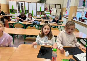 Uczniowie siedzą przy złączonych stolikach i ozdabiają gałązkami świerka obsypane cukrem słoiki.