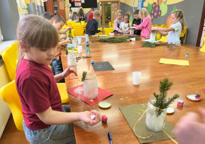 Uczniowie siedzą przy złączonych stolikach i ozdabiają gałązkami świerka obsypane cukrem słoiki.