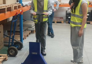 Uczeń obsługuje niebieski wózek paletowy z paletą.