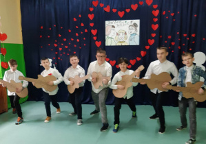 Od lewej: Bartosz, Kuba Borowiecki, Igor, Filip, Kuba Heleniak, Bartek, Michał. Chłopcy w wykroku "grają" na samodzielnie wykonanych, tekturowych gitarach.Wszyscy ubrani w białe koszule. Nad nimi plakat z narysowanymi kobietami trzech pokoleń, dookoła plakatu czerwone serca.