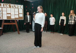 Uczennica klasy VI recytuje wiersz pt. ,,Gabryś”. Na scenie stoi tablica z hasłem ,,Julian Tuwim”. Za uczennicą recytującą wiersz stoją uczniowie, którzy biorą udział w wieczorku literackim.