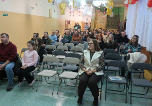 Rodzice siedzą na holu szkolnym. Są tam również uczennice i nauczyciele.