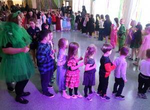 Uczniowie z nauczycielami na korytarzu szkolnym tańczą w kółku podczas balu karnawałowego.